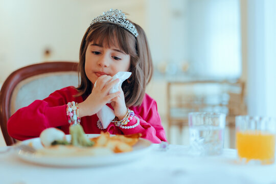 Girl with Princess Tiara Wipes Herself with Napkin at the Table. Little child being polite and respecting table manners 
