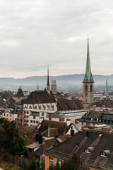 Fototapeta premium A panoramic cityscape of Zurich featuring historic church towers and rooftops under a cloudy sky. The image captures the charm of Switzerland old town with mountains in the background.