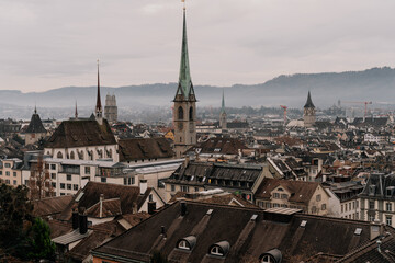 Fototapeta premium A panoramic cityscape of Zurich featuring historic church towers and rooftops under a cloudy sky. The image captures the charm of Switzerland old town with mountains in the background.