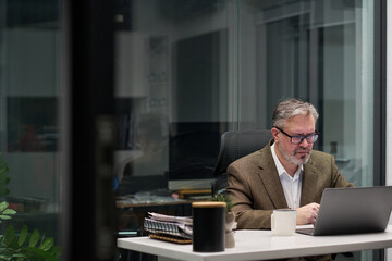 Middle aged Caucasian man working on laptop in modern office, wearing glasses and sitting at desk...