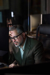 Fototapeta premium Middle aged Caucasian man wearing glasses sitting at desk working on computer, resting head on hand, appearing focused while looking at monitor in modern office setting