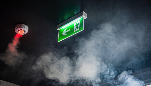 Smoke fills a hallway under an emergency exit sign with a red alarm light during an urgent situation in a building