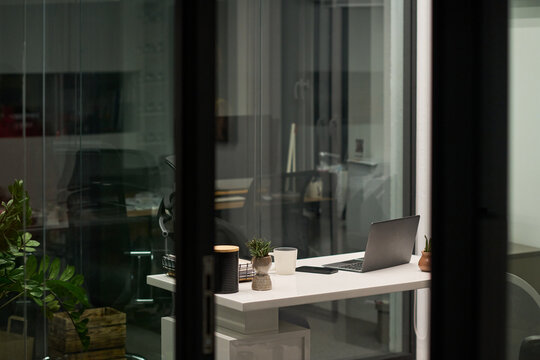 Modern office workspace featuring open laptop, coffee mug, small potted plants and stationery on desk with glass walls and empty chair, evening indoor setting visible