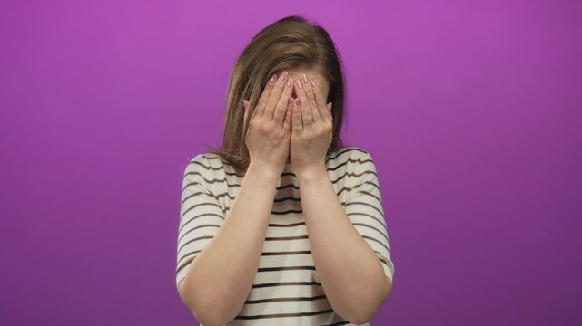 Young caucasian woman covering face with hands wearing striped shirt in purple studio; shyness vulnerability.