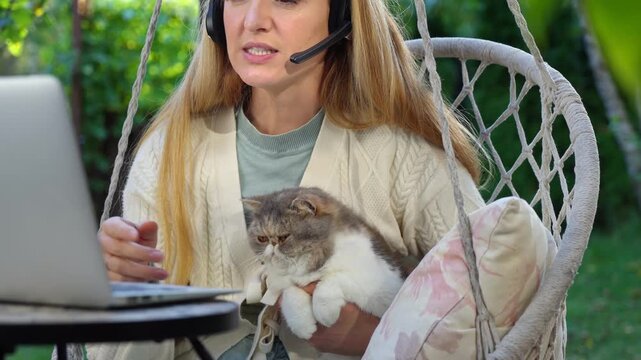 Middle aged woman working remotely with laptop and cat on her lap on terrace. Warm light, cozy lifestyle, family emotions, peaceful outdoor home office vibe.