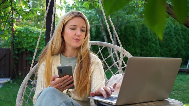 Middle aged woman working remotely on terrace with laptop and smartphone. Sunny day, cozy outdoor home office, balance of career and lifestyle, calm and focused mood.
