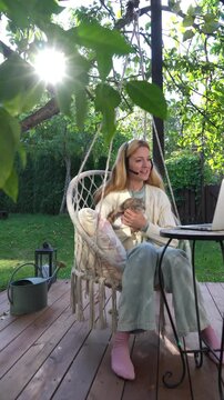 Middle aged woman working remotely with laptop and cat on her lap on terrace. Warm light, cozy lifestyle, family emotions, peaceful outdoor home office vibe.