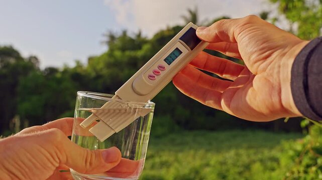 First Person View Of Hands Measuring Purity Of Drinking Water With Digital Tester Showing Twenty Ppm In Clear Glass Against Tropical Green Landscape. Clean Drinking Water Verification