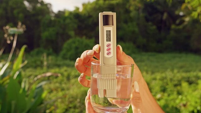 Close View Of Female Hand Holding Glass With Digital Water Quality Tester Showing Twenty Ppm Ppm Measurement Against Tropical Green Landscape. Drinking Water Safety Monitoring