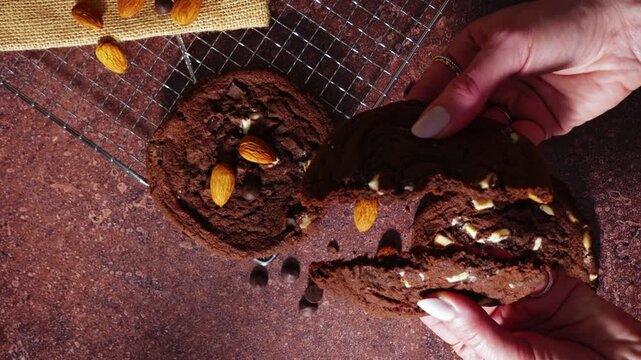 Top view of hands breaking a soft chocolate cookie with almonds and chocolate pieces. Fresh baked crumb and texture revealed on a rustic table with cooling rack and ingredients.