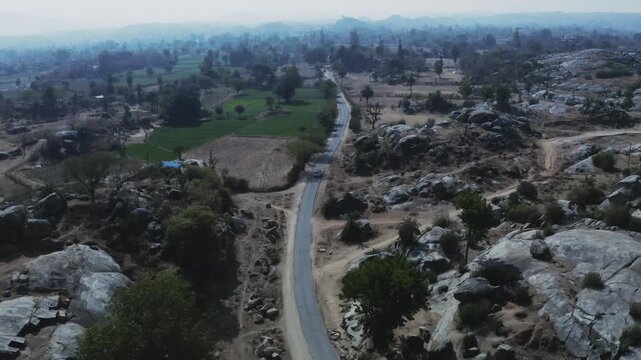 A vehicle travels a paved road cutting through a semi-arid landscape dotted with granite boulders, sparse greenery, and small farms near Rohida, Rajasthan.
