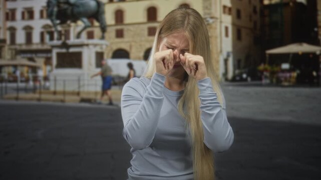 Blonde woman rubbing hands over eyes on a historic street plaza with statue and outdoor cafe in view; sadness.