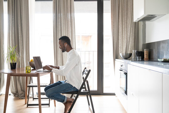 Smiling man sitting at table in modern apartment using laptop and earphones