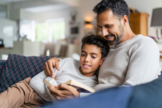 Father lying with son on couch in living room reading book