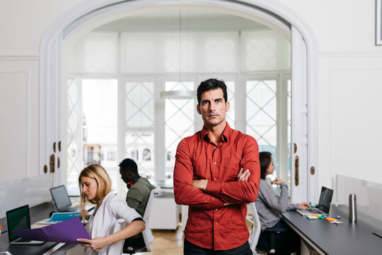 Businessman standing with arms crossed in office with colleagues working in background