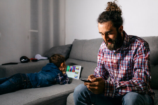 Father sitting on the couch looking at smartphone while his son using digital tablet in the background