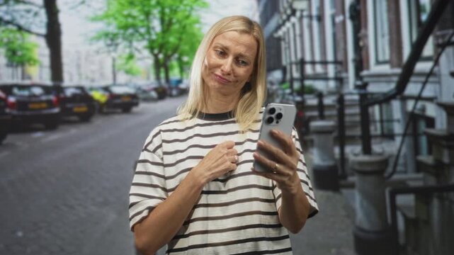 Woman holding smartphone, hand on chest, reading a message on a city street among parked cars and building stoops while wearing a striped tshirt; surprise reaction.