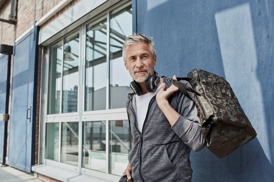 Portrait of mature man with headphones and camouflage sports bag in front of gym
