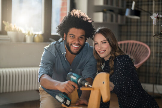 Smiling couple looking at camera mounting rocking horse with cordless drill