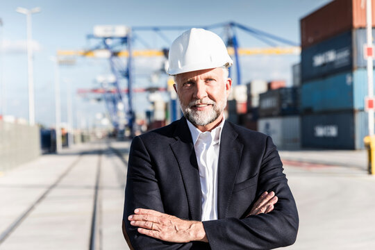 Businessman at cargo harbour, wearing safety helmet, arms crossed