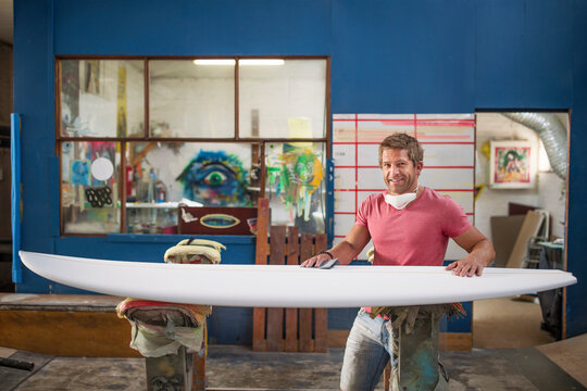 Surfboard shaper workshop, portrait of smiling surfshop employee with surfboard