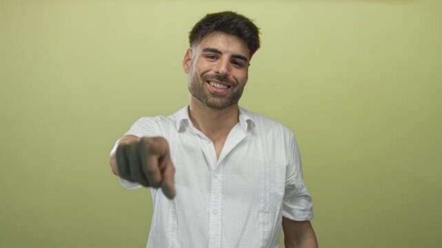Smiling young hispanic man pointing finger directly at camera while wearing a white shirt in a light green studio space; confidence.