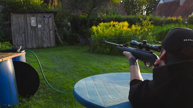 Man aiming and firing scoped rifle at a target during backyard target practice on a sunny summer day surrounded by green garden plants.