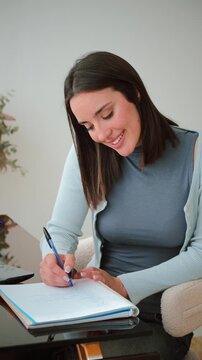 Engaged woman thoughtfully writing notes in her notebook, showcasing concentration and creativity during her work session at a stylish desk with a pen in hand and a smile on her face.