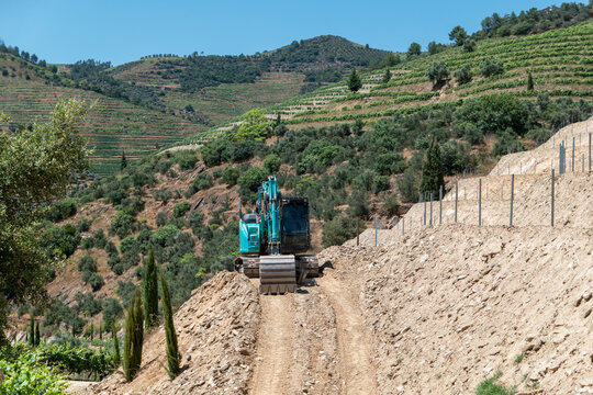 Escavadora de rastos a realizar trabalhos de terraplanagem e abertura de caminho numa encosta rural com vegeta&ccedil;&atilde;o e vinha em socalcos ao fundo, numa paisagem de montanha sob um c&eacute;u azul