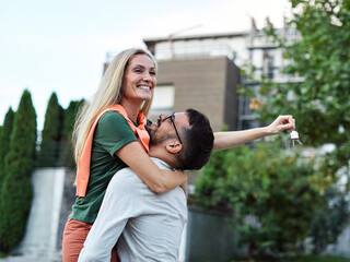 Portrait of a happy young couple standing in front of their new house, with keys in hands, smiling...