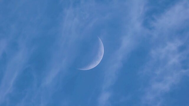 Ethereal cinematic shot of the lunar crescent surrounded by moving clouds in a blue sky.