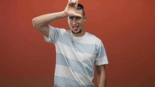 Man forms l with hand on forehead and sticks tongue out in a studio with orange backdrop wearing a striped shirt; playful mischief.