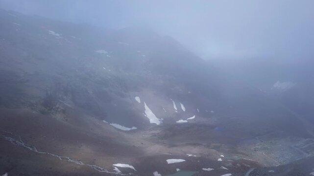 Dramatic aerial view reveals Bhoot Ground's remote high-altitude plateau on Sach Pass, surrounded by snow-patched Himalayan peaks and drifting mountain mist in Himachal Pradesh, India.