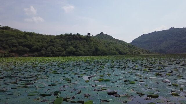 Drone glides low over lotus-covered waters of Jait Sagar Lake toward hilltop chhatri in Bundi, Rajasthan, with forested hills under overcast sky.