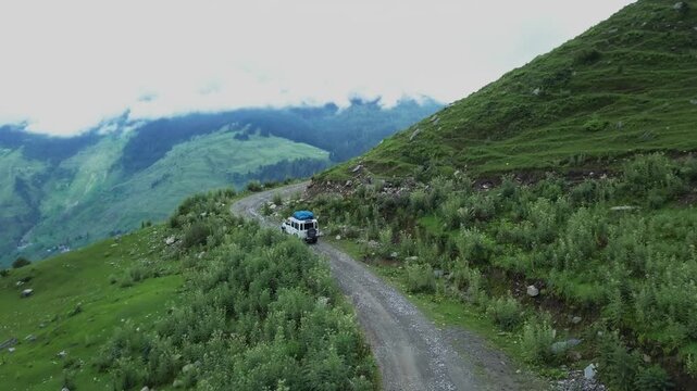 White SUV with blue roof navigates narrow dirt mountain road winding through lush green Himalayan alpine slopes with dramatic cloud-covered peaks in the distance.