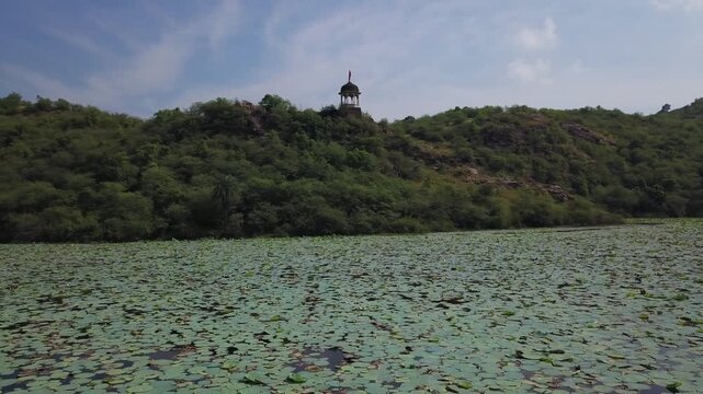 Aerial drone ascends from lotus-covered Jait Sagar Lake toward a domed pavilion on a forested hill in Bundi, Rajasthan, under a partly cloudy sky.