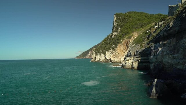 Rocky Ligurian coastline near Porto Venere facing toward Cinque Terre on a clear sunny day, with turquoise Mediterranean sea, steep cliffs, and lush green vegetation along the rugged shore.