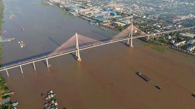 Aerial drone shot of Vam Cong Bridge crossing wide muddy Mekong River with boats moving along river channel beside urban area and industrial buildings in Vietnam