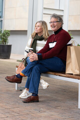 Naklejka premium Senior couple wearing sweaters and jeans sitting on wooden bench in plaza with coffee, paper bags