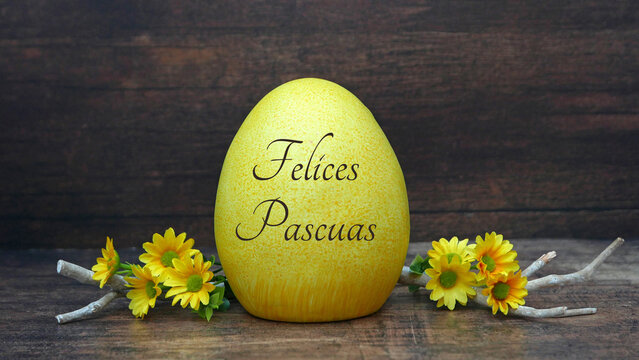 Un huevo de Pascua amarillo con la inscripci&oacute;n "Felices Pascuas" sobre una mesa de madera, rodeado de flores y ramas amarillas.