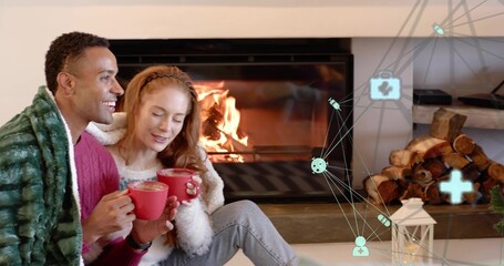 Naklejka premium Sitting couple sipping red mugs in living-room by fireplace, wearing knit sweaters, network overlay