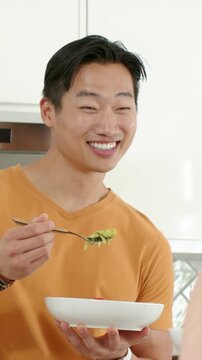 Vertical video: Seeing partner, man in orange tee scooping salad at home offering forkful to share