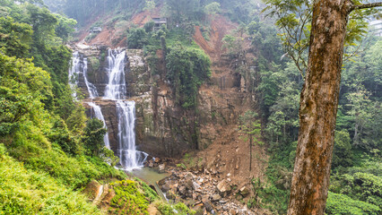 Ramboda Waterfall near Nuwara Eliya town in Sri Lanka. Majestic multi-tier waterfall cascades down a lush green mountain slope, framed by a prominent tree trunk © Viliam