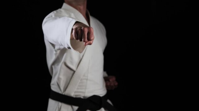 A male athlete with a black belt in karate performs punches and blocks with his hands on a black background