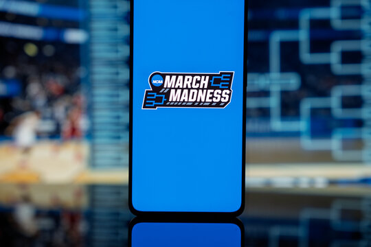 Smartphone displaying the NCAA March Madness logo on a blue screen, positioned against a blurred background of a basketball tournament bracket and arena, representing college basketball