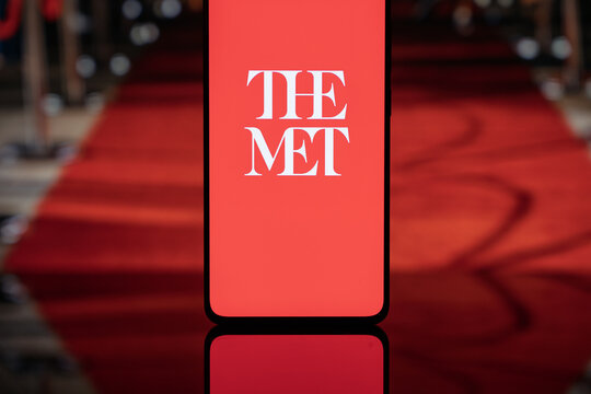 Smartphone displaying The Met logo on a red screen background, positioned in front of a blurred red carpet, representing the Met Gala, luxury fashion events, and high-society celebrity culture