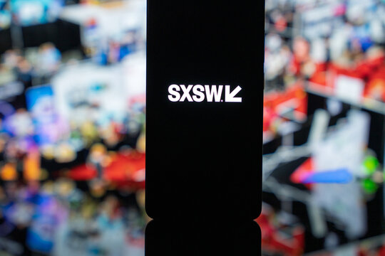 Smartphone displaying the SXSW logo against a blurred background of a busy conference or trade show floor, representing South by Southwest festival, tech innovation, and creative industry events