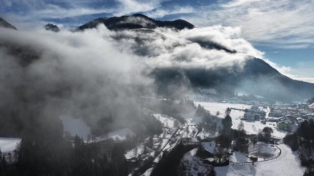 Winter aerial view of alpine village in snowy valley surrounded by mountains and clouds in the Dolomites, Trentino, Italy. Drone shot of snow covered mountain village and valley with rolling clouds