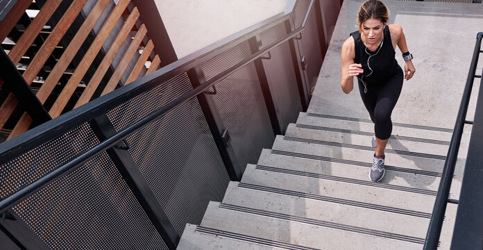 Fitness, running and woman on stairs with earphones, cardio exercise or music playlist in physical activity. Above, tech and athlete on steps with jog, podcast streaming and stamina training in city.