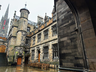 Fototapeta premium Historic stone New College Quad courtyard and gothic spire in Edinburgh, Scotland, UK with wet pavements, benches and ornate university style architecture